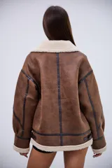 Campera tipo gamulán de corte oversize, confeccionada en cuero sintético marrón con efecto desgastado y forro interno de corderito sintético color crema. Presenta cuello solapa, cierre frontal con cremallera, detalles de tiras con hebillas en el cuello y en los laterales, y costuras reforzadas con bandas de cuero oscuro.