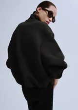 Chaqueta negra de diseño estructurado, con cuello alto, cierre frontal de cremallera metálica y mangas abullonadas con puños ajustados.