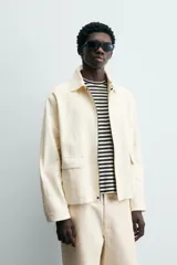 Cazadora de hombre color beige claro, corte relajado, con cuello solapa, manga larga y puños con botones a presión. Presenta bolsillos de plastrón con solapa en la cadera y cierre frontal de cremallera oculta por solapa de botones a presión.