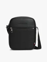 Bandolera reporter Tommy Hilfiger de color negro, confeccionada en poliuretano con acabado liso y panel frontal inferior con detalle tejido. Cuenta con un compartimento principal con cierre de cremallera, bolsillo exterior frontal también con cremallera, bolsillo interior abierto, correa ajustable y logo metálico de la marca en la parte delantera.