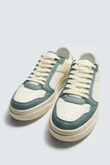 Zapatilla deportiva de estilo retro con diseño de paneles en combinación de colores crema, verde azulado y detalle en rojo en el talón. Presenta suela con volumen y cierre con cordones.