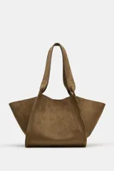 Cartera shopper de hombro confeccionada en gamuja color marrón, con diseño estructurado de laterales abiertos y doble asa superior. Cuenta con cierre de botón imantado e incluye un bolsillo interior extraíble con cremallera.