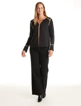 Blusa negra de crepe elastizado con ribetes en contraste color beige que recorren el escote en V, la línea central del frente y las costuras de los hombros. Tiene mangas largas con puños abiertos, abertura lateral y botón metálico.
