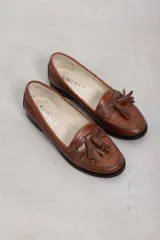 Mocasines de cuero marrón con diseño clásico, detalles de flecos y borlas en el empeine, y suela plana.