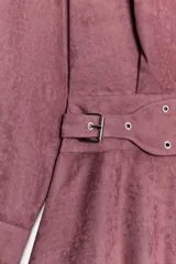 Vestido corto de cuello subido y manga larga, confeccionado en tejido efecto ante color rosado. Presenta un diseño con cinturón integrado del mismo tejido con hebilla metálica y arandelas, cierre frontal con botones ocultos y bajo asimétrico.