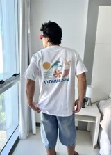 Remera blanca de manga corta con cuello redondo y corte holgado. Presenta un estampado gráfico en la espalda con elementos marinos (sol, ola, estrella de mar, flor, etc.) y el texto "VITAMIN SEA" y "BROKERS".
