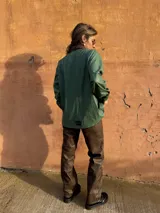 Camisa verde de cuero sintético con cuello marrón, cierre frontal con botones, dos bolsillos con solapa en el pecho y cinturón marrón.