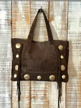 Bolso estilo shopper confeccionado en una combinación de cuero con textura croco y gamuza en color marrón. Presenta detalles de tachas metálicas circulares y flecos laterales largos, con doble asa de mano.