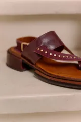 Sandalia de cuero bordó con diseño tipo thong, tiras anchas cruzadas con tachas metálicas doradas, tira con hebilla ajustable en el talón y taco bajo cuadrado con acabado artesanal.