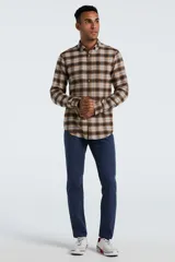 Camisa de manga larga confeccionada en tela de franela con estampado de cuadros en tonos marrones, beige y líneas azules. Presenta cuello clásico abotonado, cierre frontal con botones y un bolsillo pequeño en el pecho.