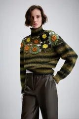 Jersey de punto con cuello subido y manga larga. Presenta un diseño de rayas horizontales en tonos verde y negro, con un panel superior decorado con calados y aplicaciones de flores tejidas en crochet de colores contrastantes.