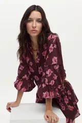 Blusa bordó con estampado floral en tonos rosados, confeccionada en algodón Swiss Dot, con mangas largas abullonadas y escote en V con botones frontales.