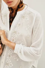Camisa blanca con cuello mao y escote en V, con bordados negros y dorados en el frente y mangas largas con puños abotonados.