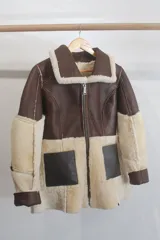 Gamulán reversible de cuero genuino color marrón oscuro con interior de piel sintética en tono crema. Presenta cuello solapa, cierre frontal con cremallera y mangas largas con puños de piel.