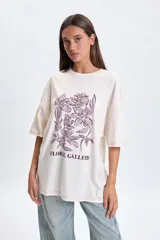 Remera de corte oversize color crudo, con cuello redondo y mangas cortas. Presenta una estampa frontal de flores en tonos violeta y el texto 'FLORAL GALLERY' en la parte inferior.