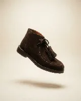 Bota de caña media confeccionada en cuero gamuzado color marrón chocolate. Presenta un diseño de estilo mocasín con cordones frontales decorados con borlas desmontables y suela plana de goma resistente.