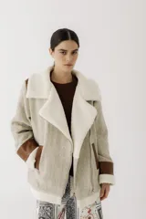 Chaqueta tipo gamulán confeccionada en paño con textura de espiga en tono beige jaspeado. Presenta solapas amplias, puños y bordes inferiores forrados en corderito sintético color crema. Incluye detalles en cuero sintético marrón en los bolsillos y puños, con cierre frontal cruzado.