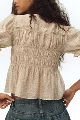 Blusa de lyocell con estampado floreado pequeño sobre base beige, escote redondo con tiras decorativas para atar, busto fruncido, mangas cortas abullonadas con elástico y ribete de volado, y bajo levemente acampanado.