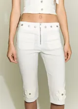 Pantalon capri blanco de tiro alto con diseño entallado. Presenta cierre frontal con cremallera, botones decorativos en la cintura, dos bolsillos de parche delanteros y detalle de flores aplicadas en la parte inferior de cada pierna.
