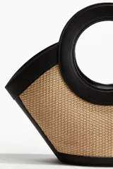 Cartera de mano con cuerpo de paja trenzada y detalles en cuero sintético negro. Presenta un diseño estructurado con asas circulares integradas y cierre magnético.