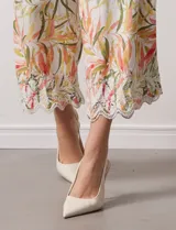 Pantalón capri de lino color crudo con estampado de hojas en tonos verdes, amarillos y naranjas. Presenta bolsillos laterales y detalle de puntilla en el ruedo.