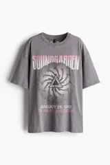 Remera de algodón gris oscuro con efecto desgastado, corte oversized y hombros caídos. Estampado frontal con el nombre de la banda "Soundgarden" en rosa pálido, un gráfico circular con un triángulo en el centro y la fecha y lugar de un concierto: "January 29, 1992 The Palace Hollywood".