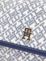 Cartera tipo mensajera con solapa, confeccionada en poliuretano con estampado integral del monograma TH. Presenta detalles en color azul marino, herrajes dorados y el logo metálico TH en el frente. Incluye asa superior extraíble y cierre a presión.