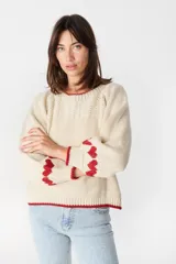 Sweater de punto color crudo con diseño de corazones rojos en las mangas y detalles de ribete rojo en cuello, puños y ruedo. Presenta un corte holgado con mangas abullonadas.