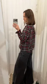 Camisa de algodón a cuadros con patrón en tonos rojo, negro y gris. Presenta cuello alto con volados, mangas largas con puños fruncidos y cierre frontal con botones.