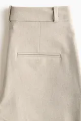 Pantalón sastrero de sarga color beige, con cintura clásica, trabillas para cinto y cierre frontal oculto. Presenta bolsillos laterales al bies, pliegues frontales y piernas rectas con raya marcada.