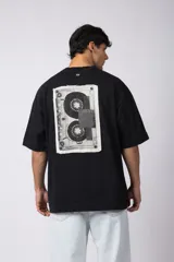 Remera negra de corte oversize confeccionada en tela pesada. Presenta un bordado frontal con la palabra 'REWIND' y un diseño de parche en la espalda con estética de papel roto.