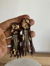 Caravanas artesanales con base superior de resina en color marrón con detalles de lámina dorada y cristales incrustados. Presentan flecos largos de hilo marrón combinados con tiras de strass brillante.