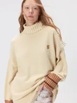 Pullover de cuello tortuga en color beige, con diseño holgado, puños acanalados en contraste y logo bordado en el pecho.
