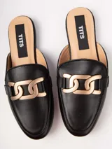 Zueco tipo mule negro, con puntera redonda y adorno de hebilla metálica dorada entrelazada.