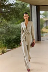 Blazer corto de satén color beige dorado, con escote en V profundo, hombreras y un lazo grande integrado en la cintura para anudar al frente. Presenta mangas largas de corte recto.
