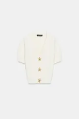 Chaqueta corta de punto texturizado color blanco roto, con escote en V, manga corta y cierre frontal con tres botones metálicos dorados en forma de estrella de mar.