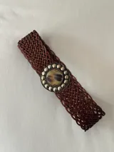 Cinto ancho de cuero trenzado en color marrón, con hebilla circular decorativa que presenta un centro de piedra natural y borde con apliques metálicos esféricos.