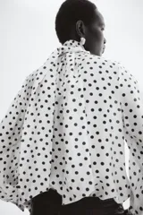 Blusa de gasa color beige con estampado de lunares negros, cuello alto con moña integrada y mangas largas abullonadas con puños ajustados.