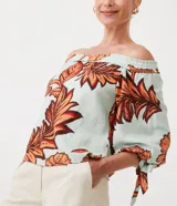 Blusa celeste con estampado de hojas color naranja y marrón, con escote hombro a hombro, mangas 3/4 con lazos en los puños y corte holgado.