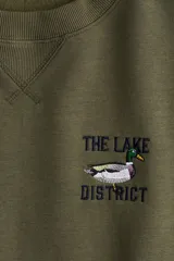 Buzo oversized color verde militar con interior cepillado suave. Presenta hombros caídos y un diseño de texto bordado en el pecho que dice "THE LAKE DISTRICT" con un pato. Tiene ribete acanalado en cuello, puños y bajo.