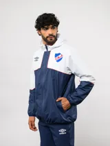 Campera de lluvia Umbro del Club Nacional de Football, con diseño de bloques de color en rojo y azul marino. Presenta capucha, cierre frontal, puños elásticos y detalles de piping blanco. Incluye logo de Umbro y escudo del club en el pecho.