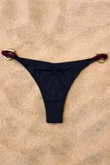 Bombacha de bikini negra con textura acanalada, presenta detalles de estribos metálicos dorados en los laterales unidos por cordones en tono bordó.