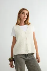 Chaleco de punto con escote en V y cierre frontal de botones. Presenta un diseño de corte recto y textura jaspeada en tono crema.