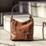 Cartera estilo hobo color marrón, confeccionada en material símil cuero con textura. Presenta dos bolsillos frontales con cierre metálico, apertura superior con cierre y asa corta para el hombro.