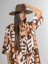 Vestido largo de manga tres cuartos, con cuello camisero y botones frontales. Presenta un estampado abstracto en tonos marrón oscuro, beige y blanco. El calce es amplio y tiene un ruedo con volado.