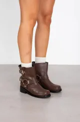 Botas de caña baja en ecocuero color marrón, con diseño de estilo biker que incluye dos correas con hebillas metálicas y una argolla decorativa en el tobillo. Posee suela y taco bajo de color negro.