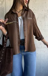 Camisa de gamuza color chocolate con bordados decorativos en color crudo a lo largo de la botonadura frontal y en el bolsillo superior. Presenta cuello clásico, cierre frontal con botones y mangas largas.