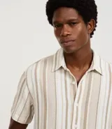 Camisa de hombre de manga corta con cuello tradicional y abotonado frontal. Presenta un tejido texturizado con un patrón de rayas verticales en tonos beige, blanco y marrón claro.