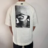 Remera oversize de algodón pesado color crema o blanco roto, con un gran estampado fotográfico en blanco y negro en la espalda que muestra la boca de una persona fumando un cigarrillo, con un gorro tejido cubriendo parcialmente el rostro.