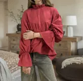 Blusa de algodón color bordó, con cuello alto estilo victoriano con volados, mangas largas abullonadas con puños elásticos y corte holgado con detalle de costura horizontal en el pecho.